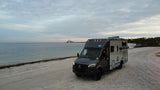 An RV rental, a Winnebago EKO, rests on a sandy beach. A bridge spans the bay under a cloudy sky. Perfect for waterfront travel.