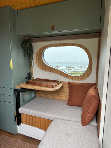 Cozy RV rental interior with a unique window framing an ocean view, light seating, green cabinets, and a pull-out table.