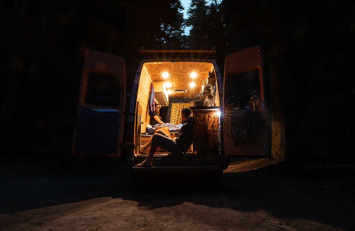 Two people relax inside a brightly lit RV rental camper van at night, doors open to nature.