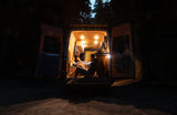 Two people relax inside a brightly lit RV rental camper van at night, doors open to nature.