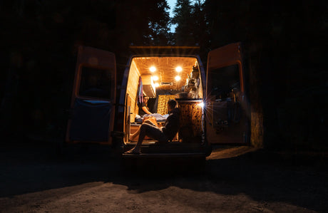 Two people relax inside a brightly lit RV rental camper van at night, doors open to nature.