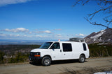 White rv rental van with rooftop tent parked on a mountain road, overlooking a vast forest and distant water under blue sky.