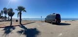 Black Sprinter RV rental parked by the ocean with palm trees and a pier under a clear sky.