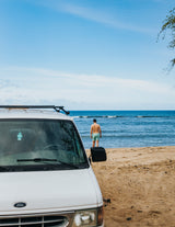 White Ford camper van (rv rental) on a sandy beach with a man standing in the ocean facing calm waves.