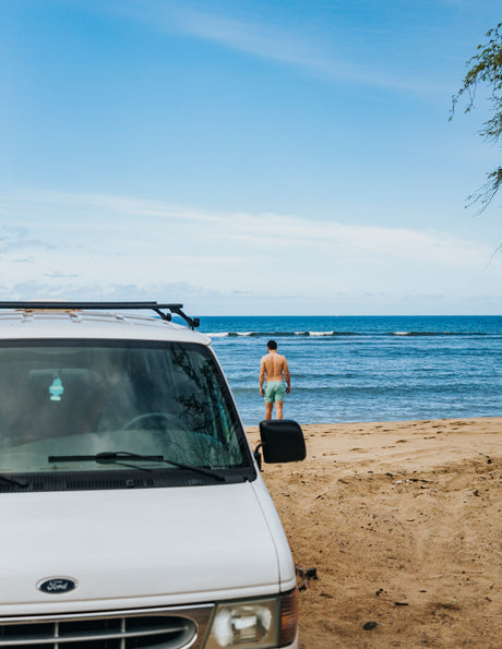 White Ford camper van (rv rental) on a sandy beach with a man standing in the ocean facing calm waves.