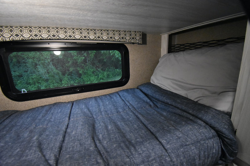 Cozy RV rental sleeping area with a window offering a lush green view of nature. Comfortable bed for travel adventures. — photo 13