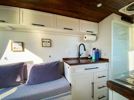 Bright RV rental interior features a mini kitchen with sink, white cabinets, wood ceiling, and cozy seating.