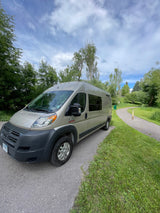 Tan Ram ProMaster RV rental parked on a scenic paved path surrounded by green trees, ideal for a road trip.