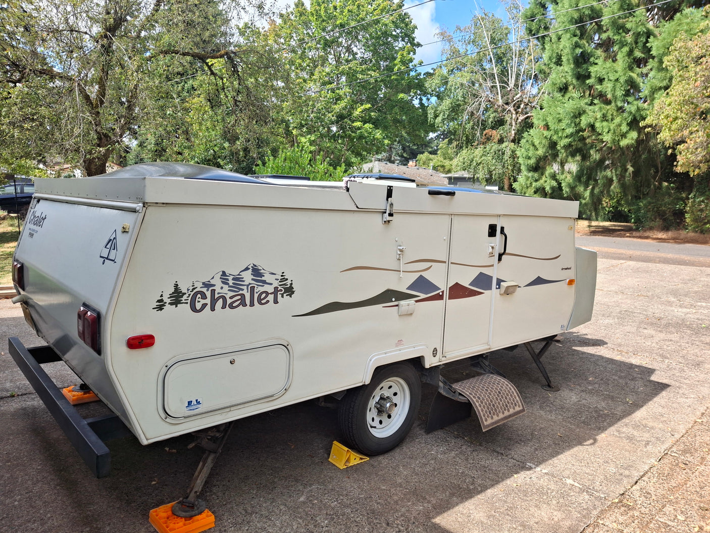 White Chalet pop-up camper, perfect for an RV rental. Parked on concrete with stabilizing jacks and mountain decals.