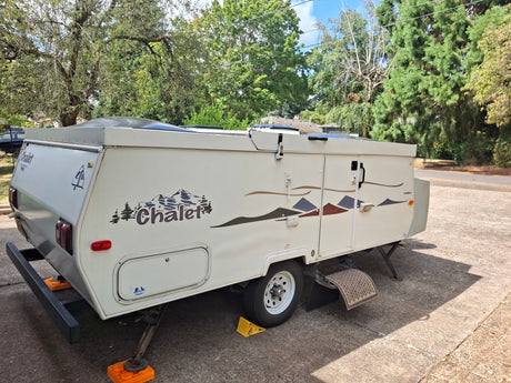 White Chalet pop-up camper, perfect for an RV rental. Parked on concrete with stabilizing jacks and mountain decals.