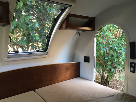 Cozy rv rental interior featuring a bed, wood paneling, a camper window, and an open archway showcasing green trees.