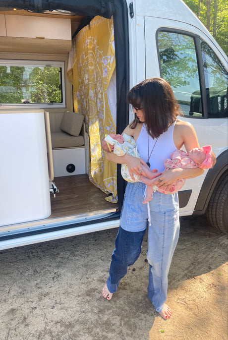 A woman holding newborn twins outside a white RV rental; its cozy interior with yellow patterned curtains visible, perfect for family travel.