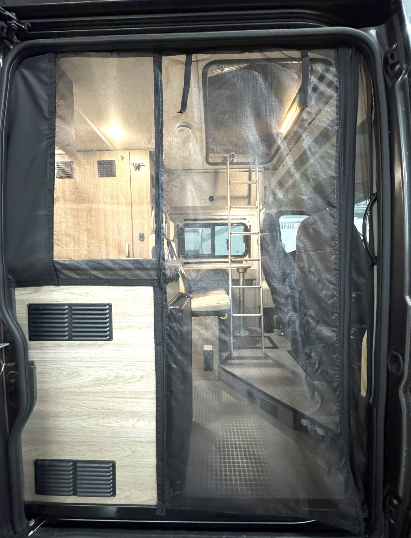 Step inside this **RV rental**! View of a camper van interior through a screen door, showing a bed, ladder, and dinette area.