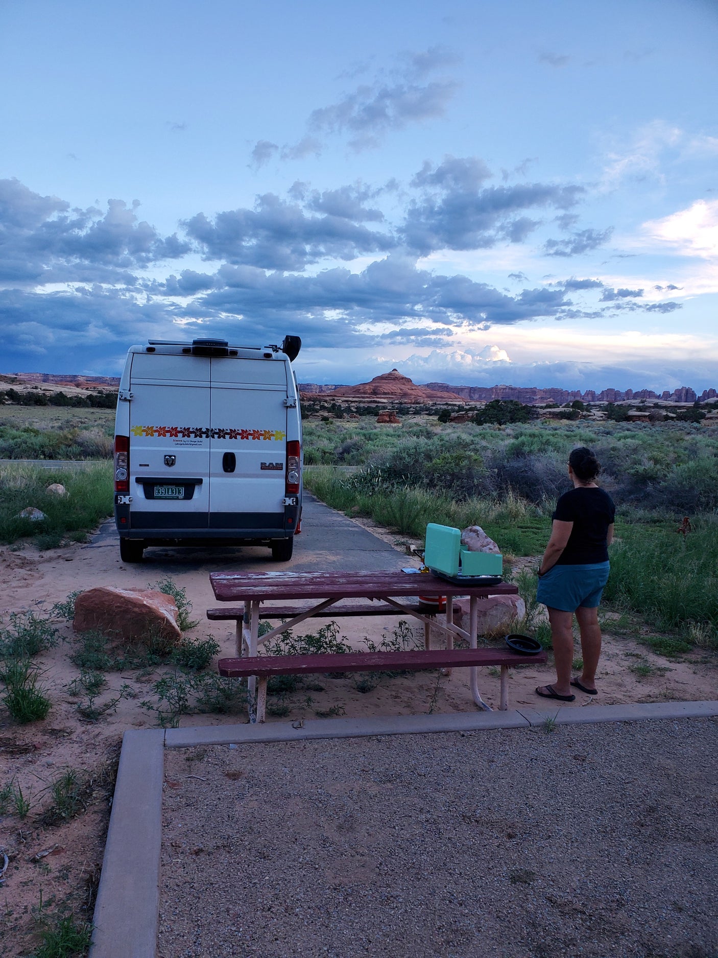 Evening at a desert campsite. An RV rental is parked while a person admires the red rock landscape under a cloudy sky.