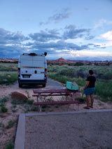 Evening at a desert campsite. An RV rental is parked while a person admires the red rock landscape under a cloudy sky.