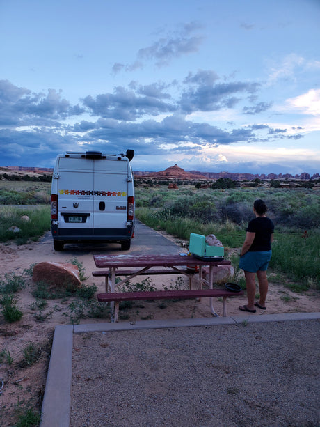 Evening at a desert campsite. An RV rental is parked while a person admires the red rock landscape under a cloudy sky.