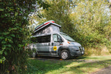 A grey camper van RV rental with a pop-top roof and paddleboards is parked amidst lush trees at a sunny campsite.