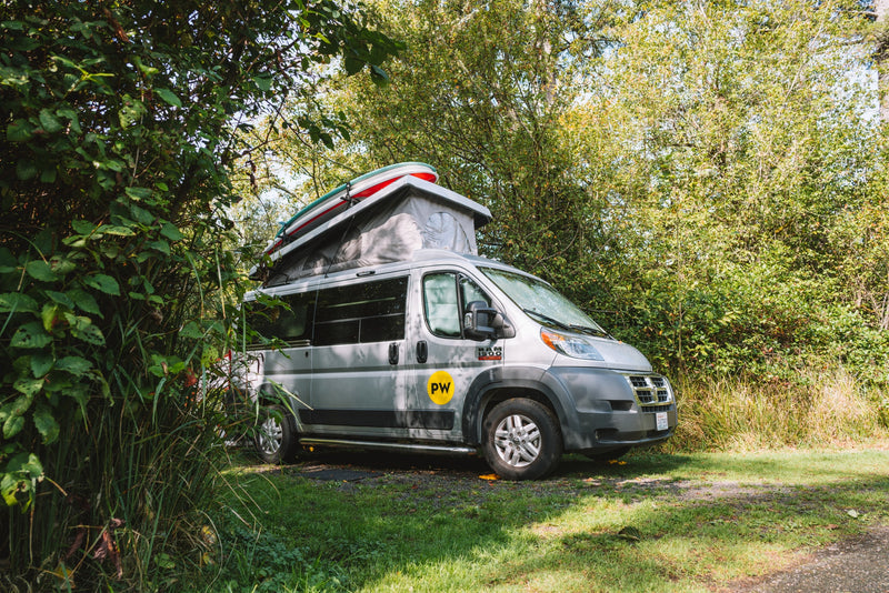 A grey camper van RV rental with a pop-top roof and paddleboards is parked amidst lush trees at a sunny campsite. — photo 6