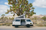 White RV rental camper van with open pop-top roof parked among Joshua trees in a vast desert landscape.