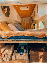 Cozy RV rental interior showcasing a comfortable bed, overhead cabinets, and clever under-bed storage with firewood.