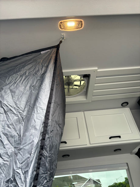 Interior of an **rv rental** showing a ceiling light, fan, and a hook holding a dark gray privacy curtain. White cabinets below.