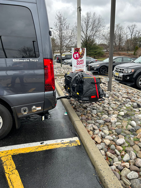 This 'Ultimate Toys' RV rental shows its rear, loaded with cargo on a carrier in a wet parking lot next to a donut ad.