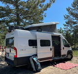 White Winnebago Solis RV rental set up for camping under pine trees. Pop-top roof is open, side door open to interior.