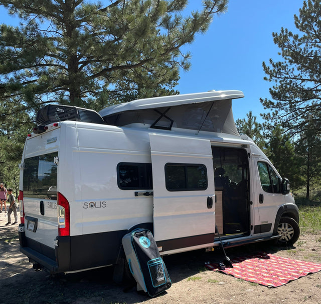 White Winnebago Solis RV rental set up for camping under pine trees. Pop-top roof is open, side door open to interior.