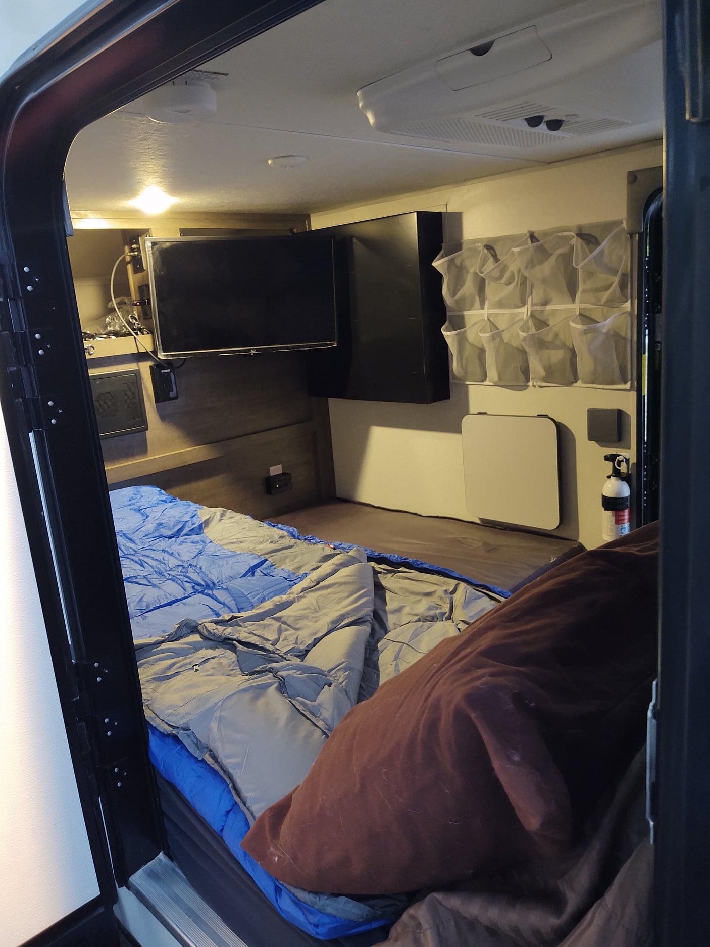 Cozy RV rental interior: bed with sleeping bag, TV, hanging storage, small table, and a wall-mounted fire extinguisher for safety.