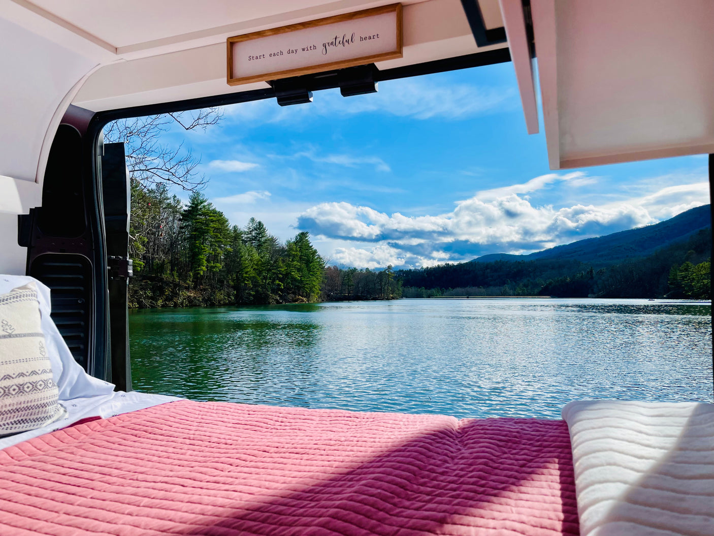 Cozy RV rental bed with an expansive view of a serene lake, lush trees, and majestic mountains. Embrace van life.