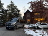 Experience winter! This RV rental, a Mercedes camper van, is parked at a snowy log cabin with evergreens.