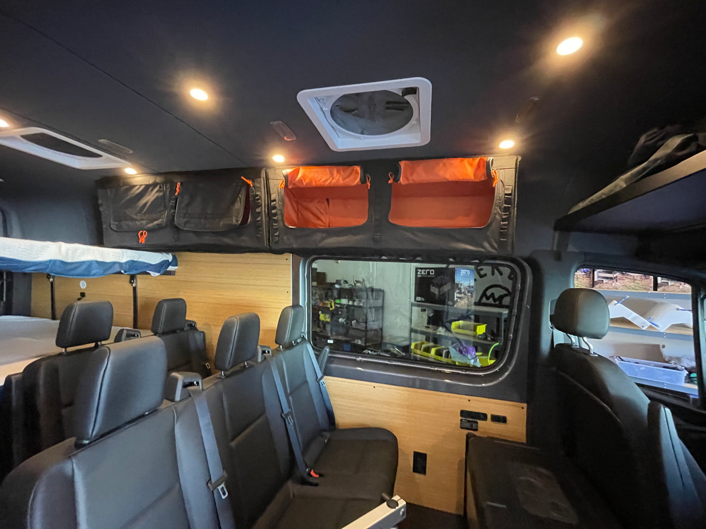 Inside an RV rental: elevated bed, passenger seating, overhead storage (black/orange bags), wood paneling, ceiling fan.