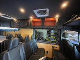 Inside an RV rental: elevated bed, passenger seating, overhead storage (black/orange bags), wood paneling, ceiling fan.