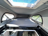 RV rental interior: A raised bed platform, overhead skylight with blue mesh, and side windows in a pop-top camper.