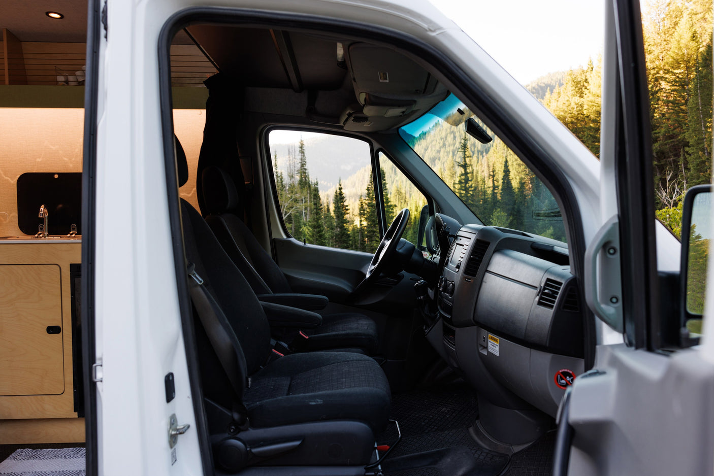 This RV rental showcases its cabin & kitchenette, with a driver's perspective of scenic mountains & a vibrant forest outside.