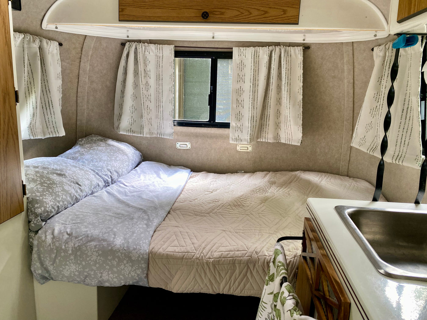 Cozy rv rental interior: comfortable bed with floral and quilted bedding, windows with curtains, and a compact sink. — photo 8