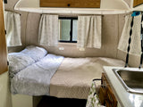 Cozy rv rental interior: comfortable bed with floral and quilted bedding, windows with curtains, and a compact sink.
