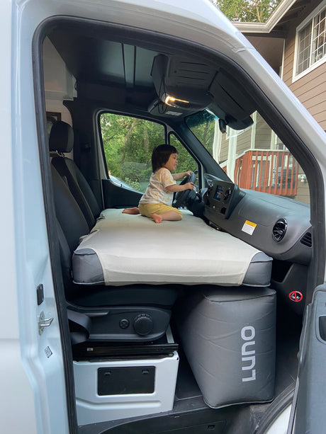 Inside an RV rental van, a child sits on a front cab bed (likely Luno brand) playing with the steering wheel.