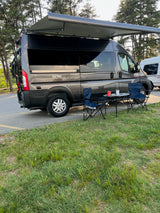 A grey campervan RV rental is parked with its awning extended, featuring an outdoor setup with chairs and a table at a campsite.