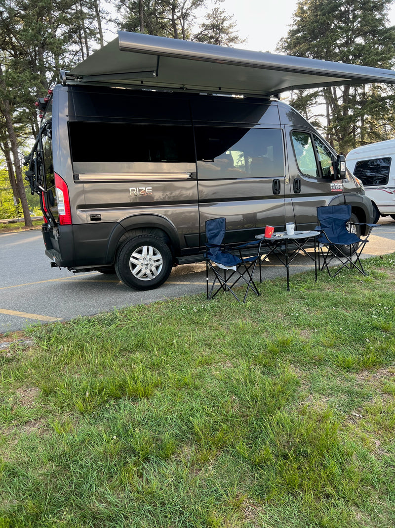A grey campervan RV rental is parked with its awning extended, featuring an outdoor setup with chairs and a table at a campsite. — photo 8