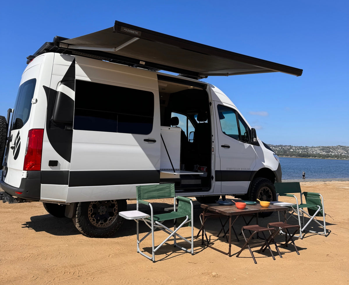 Enjoy the perfect beach day with this RV rental! A Storyteller Overland camper van with a Dometic awning set up on sand by water.