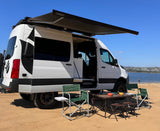 Enjoy the perfect beach day with this RV rental! A Storyteller Overland camper van with a Dometic awning set up on sand by water.