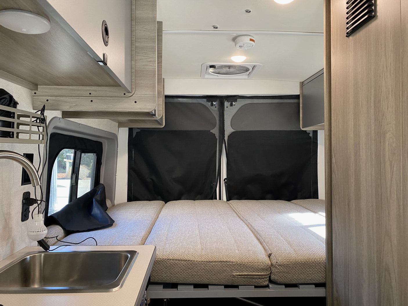 Inside this compact RV rental: a spacious bed area with black window covers, plus a mini kitchen sink & overhead storage.