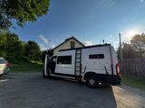 White rv rental camper van with open door and ladder, parked on gravel. Sunny day, house and trees visible in background.