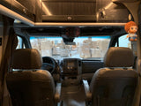 Inside an RV rental: brown front seats and dashboard facing stacks of boxes in a warehouse. A Bob Ross doll hangs.