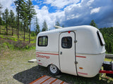 White Scamp RV rental parked in a beautiful, tree-lined campsite under a partly cloudy sky. Ideal for nature lovers.