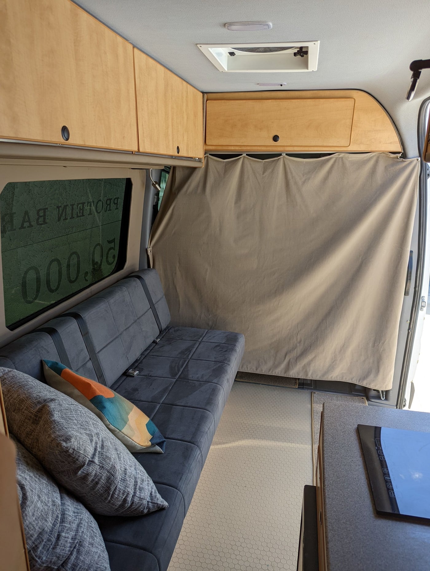 Comfortable RV rental interior with grey sofa, overhead wood cabinets, tiled floor, and a rear privacy curtain. — photo 11