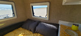 Cozy RV rental interior with a stunning beach view. Yellow bedding, grey pillows, and ocean scenery through the windows.