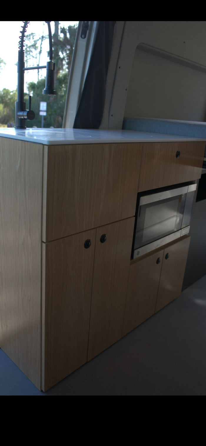 Modern rv rental kitchen with light wood cabinets, white counter, built-in microwave, and sleek black faucet inside a camper van.