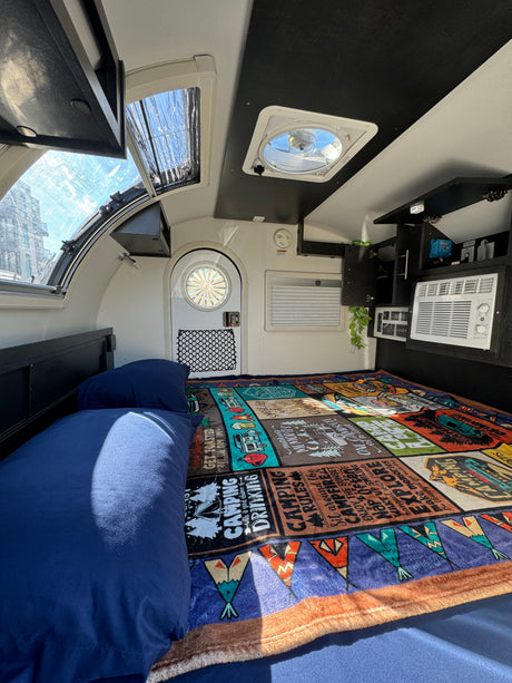 Cozy RV rental bedroom with a colorful camping blanket, blue pillows, AC unit, and ceiling vents.